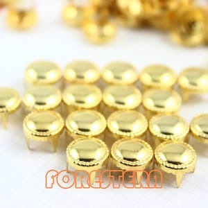 May include: A close-up of a group of gold-colored metal studs with a decorative edge. The studs have three prongs on the bottom for attaching to fabric.