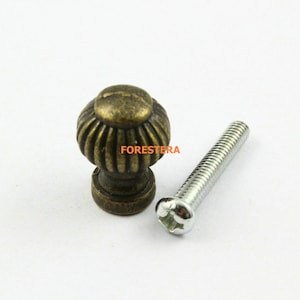 May include: A close-up of a small, antique-style cabinet knob with a fluted design. The knob is made of metal and has a bronze finish. It comes with a silver screw.