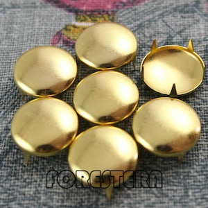 May include: Eight gold-colored metal round buttons with a raised center and a pointed base. The buttons are arranged in a circle on a textured fabric surface.