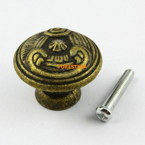 May include: A gold-toned metal cabinet knob with an ornate design and a matching screw. The knob has a raised, circular pattern with a floral motif.