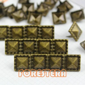 May include: A close-up of a set of antique gold pyramid-shaped studs with a decorative border. The studs are arranged in rows on a white surface.