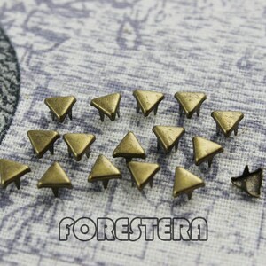 May include: A collection of 18 gold-toned metal triangle studs with three prongs on the back. The studs are scattered on a textured fabric surface.