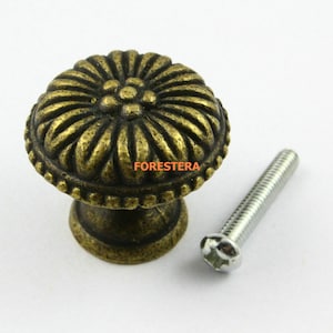 May include: Antique brass cabinet knob with a floral design and a matching screw. The knob has a raised, textured center with a smaller flower design.