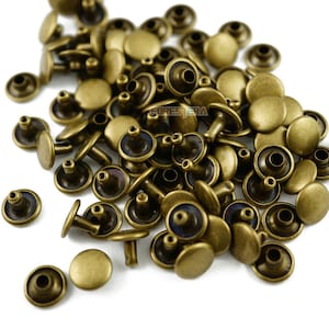 May include: A pile of antique brass-colored rivets. Each rivet has a flat, round head and a post on the back. The rivets are scattered across a white surface, with some overlapping. The rivets are likely used for leathercraft or garment construction.