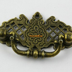 May include: Antique brass-toned drawer pull with an ornate, openwork design. The pull features a decorative handle and two spherical end pieces. The center of the pull has a floral design. The overall shape is a stylized, symmetrical design.
