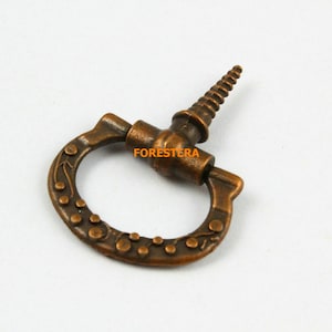 Pode incluir: Uma puxador de gaveta de metal de cor bronze, de estilo antigo e decorativo, com um verso de parafuso. A puxador tem um design circular e ornamentado com um padrão floral e pequenos pontos.