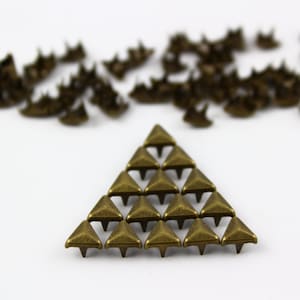 May include: A group of small, gold-toned triangle-shaped metal studs with pointed tips. The studs are arranged in a triangle shape.