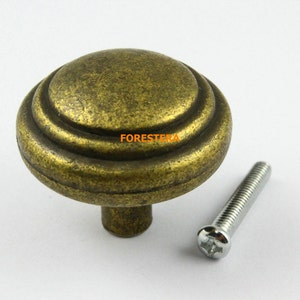 Könnte beinhalten: Ein runder, antiker goldfarbener Schrank-Knopf mit drei konzentrischen Ringen und einer Schraube. Der Knopf hat eine patinierte Oberfläche.