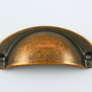 Op de afbeelding: Een koperkleurige metalen kastgreep met een afgeronde, gebogen vorm. De greep heeft twee gaten voor bevestiging.