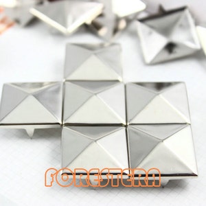 Könnte beinhalten: Ein Satz silberner pyramidenförmiger Nieten, die in einem Kreuzmuster angeordnet sind. Die Nieten haben eine glänzende, metallische Oberfläche und sind für die Verwendung in Bastelarbeiten oder DIY-Projekten gedacht.