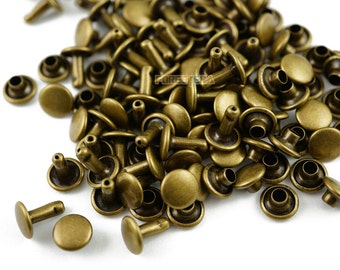 May include: A pile of antique gold metal rivets. The rivets have a round head and a post with a hole in the center. The rivets are used for attaching fabric, leather, or other materials.
