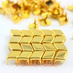 May include: Close-up of gold-colored metal diamond-shaped studs, arranged in a grid pattern. Additional studs are scattered in the background. The studs have a shiny, reflective surface. The word "FORESTERA" is printed in orange.