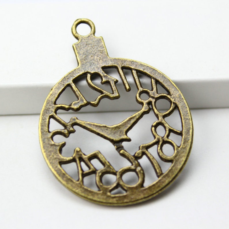 May include: A bronze-colored metal charm shaped like a clock face with Roman numerals and a cutout design. The charm has a loop at the top for attaching to a necklace or bracelet.