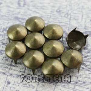 May include: A group of 10 antique gold-toned metal pyramid-shaped studs with a pointed tip. The studs are arranged in a semi-circle on a white fabric background.