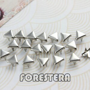 May include: A collection of silver triangle-shaped metal studs, scattered on a white background with a pattern of concentric circles.
