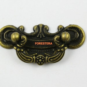Puede incluir: Un tirador de cajón de metal dorado con un diseño decorativo y ornamentado. El tirador tiene dos pomos redondeados a cada lado y un diseño central ornamentado.