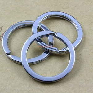 May include: Three shiny, silver-colored key rings are interlocked, showcasing a reflective surface. The key rings are circular with a hinged opening, designed for attaching keys or other small items. The background is a light brown color.