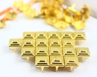 Puede incluir: Un grupo de 25 tachuelas de metal de color dorado en forma de pirámide con base cuadrada. Las tachuelas están dispuestas en un patrón rectangular.
