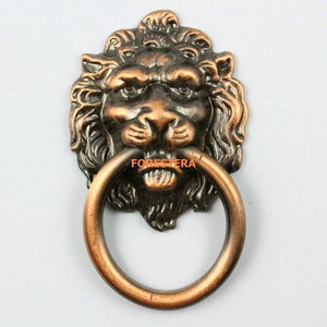 Peut inclure: Un heurtoir de porte en bronze en forme de tête de lion avec une poignée en anneau. Le visage du lion est détaillé avec une crinière et une expression féroce.