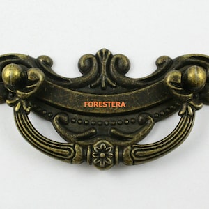 Peut inclure: Poignée de tiroir en laiton de style antique avec un motif floral décoratif et une poignée incurvée. La poignée a une finition vieillie et le mot "FORESTER" est visible sur le devant.