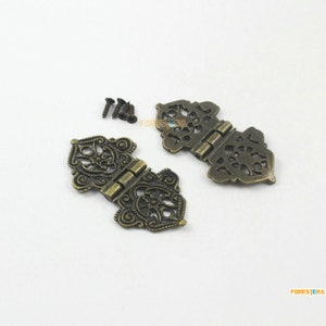 May include: Two antique-style brass hinges with intricate floral designs. The hinges are closed and have a decorative, ornate look. They come with four small screws.
