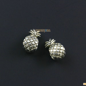 Puede incluir: Dos abalorios de piña plateados. Los abalorios están detallados con un diseño de piña texturizado y un pequeño tallo en la parte superior.