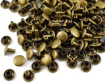May include: A large pile of antique brass rivets, with a few scattered on the white surface. The rivets have a round head and a short, straight shank.