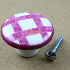 Peut inclure: Bouton de meuble en céramique à carreaux blancs et magenta avec une base blanche assortie. Une vis argentée est positionnée à droite du bouton. Le mot "FORESTERA" est imprimé sur le bouton.