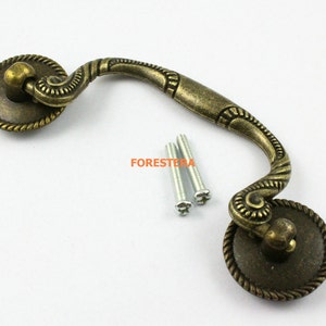 May include: Antique brass cabinet pull with ornate details and a curved design. The pull has two round knobs with rope-like edging. Two screws are included.