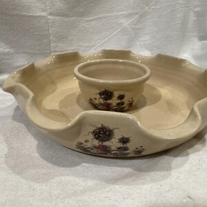 May include: A cream-colored ceramic chip and dip serving set featuring a large, round, ruffled-edge platter with a smaller, matching bowl centered inside. Both pieces are decorated with a detailed floral print featuring dark, muted botanical motifs.
