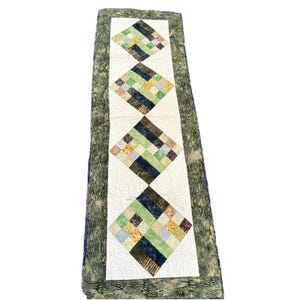 May include: A rectangular table runner featuring a white central panel with four diamond-shaped patchwork blocks. Each block is composed of small squares in shades of green, blue, yellow, and brown. The runner has a wide border with a green wood grain pattern.
