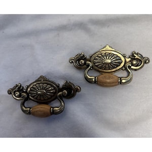 Op de afbeelding: Twee antieke messing lade grepen met een bloemen design en houten handvatten. De grepen hebben een sierlijke, vintage stijl, perfect om een vleugje elegantie aan meubels toe te voegen. Elke greep is ongeveer 13 cm lang.