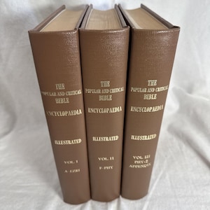 May include: Three brown hardback books titled The Popular and Critical Bible Encyclopaedia, Illustrated. The spines are labelled Vol I A-Ezri, Vol II F-Phy, and Vol III Phy-Z Appendix in gold lettering. These books feature a textured finish.
