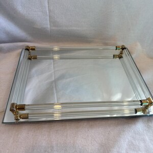 May include: A rectangular mirrored tray with clear acrylic handles and gold-toned metal corner accents. The tray has a reflective surface and is designed for decorative or serving purposes.