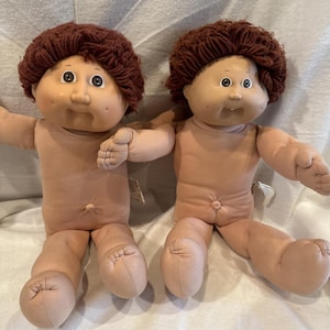 May include: Two vintage Cabbage Patch Kids dolls. Each doll has a soft, flesh-toned body, a round face with painted features, and a brown yarn hairstyle. The dolls are sitting side-by-side, with their arms outstretched.