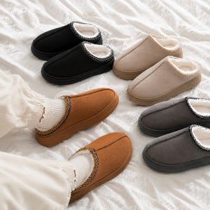 May include: A collection of slip-on slippers in black, beige, brown, and dark gray. Each slipper features a rounded toe, a thick rubber sole, and a white faux-fur lining with a decorative stitched trim along the collar. One pair is worn on feet.