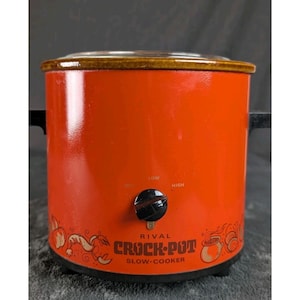 Pode incluir: Uma panela de cozimento lento Rival Crock-Pot laranja com tampa de vidro e alças pretas. O botão de controle é preto e os textos "LOW" e "HIGH" são visíveis. A Crock-Pot tem uma borda marrom e gráficos decorativos.