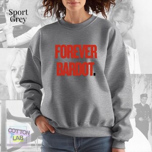 May include: Sport grey sweatshirt with the text "FOREVER BARDOT" in red. The sweatshirt is a neutral color and has a crew neck. The text is in a bold, sans-serif font. The background features a collage of images.