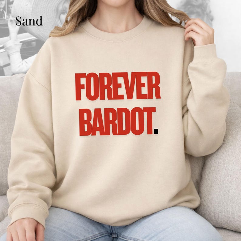 May include: Sand-colored sweatshirt with the text "FOREVER BARDOT" in bold red letters. The sweatshirt has a crew neck and long sleeves. The text is centered on the front of the shirt.