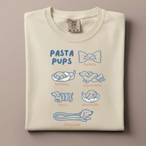 May include: Cream-colored sweatshirt with a blue graphic that reads "PASTA PUPS" and features illustrations of different pasta shapes as dogs: farfalle, tortellini, pappardelle, rotini, orecchiette, and linguine. The sweatshirt has a crew neck and a tag that reads "COMFORT COLORS".