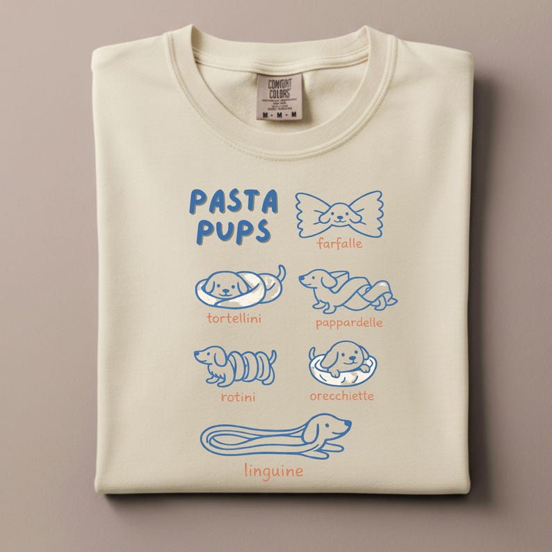May include: Cream-colored sweatshirt with a blue graphic that reads "PASTA PUPS" and features illustrations of different pasta shapes as dogs: farfalle, tortellini, pappardelle, rotini, orecchiette, and linguine. The sweatshirt has a crew neck and a tag that reads "COMFORT COLORS".