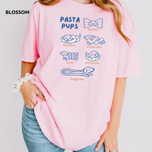 May include: A light pink t-shirt with the text "PASTA PUPS" and illustrations of pasta shapes as dogs. The shirt features drawings of farfalle, tortellini, pappardelle, rotini, orecchiette, and linguine. The word "BLOSSOM" is in the top left corner.