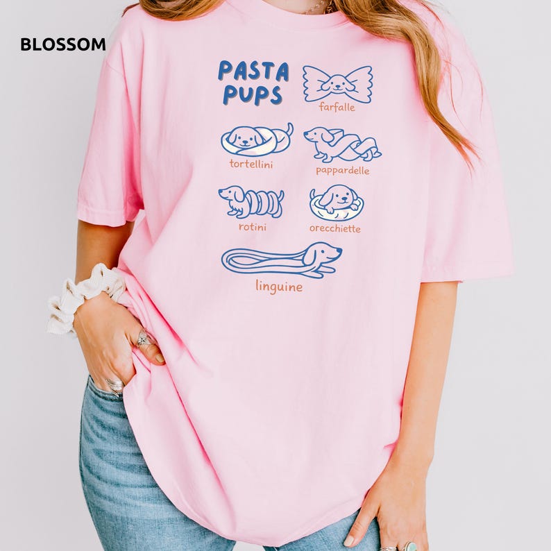 May include: A light pink t-shirt with the text "PASTA PUPS" and illustrations of pasta shapes as dogs. The shirt features drawings of farfalle, tortellini, pappardelle, rotini, orecchiette, and linguine. The word "BLOSSOM" is in the top left corner.