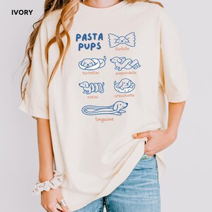 May include: Ivory-colored t-shirt with the text "PASTA PUPS" in blue, featuring illustrations of pasta shapes with dog faces. The pasta shapes include farfalle, tortellini, pappardelle, rotini, orecchiette, and linguine.