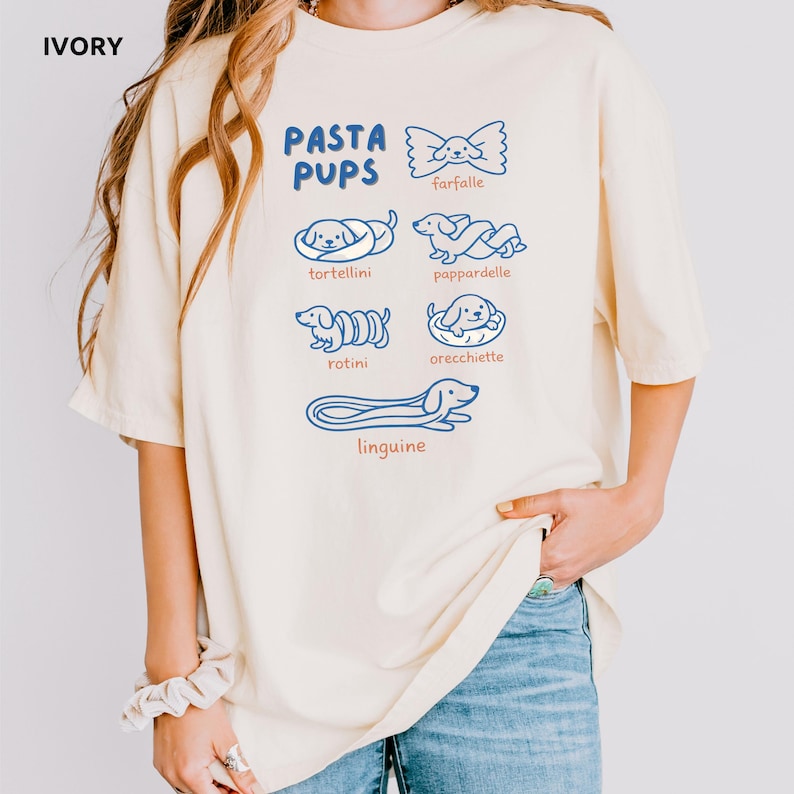May include: Ivory-colored t-shirt with the text "PASTA PUPS" in blue, featuring illustrations of pasta shapes with dog faces. The pasta shapes include farfalle, tortellini, pappardelle, rotini, orecchiette, and linguine.
