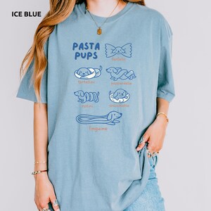 May include: Ice blue t-shirt with the text "PASTA PUPS" and illustrations of pasta shapes with dog faces. The shirt features drawings of farfalle, tortellini, pappardelle, rotini, orecchiette, and linguine.