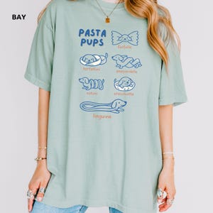 May include: A light green t-shirt with the text "PASTA PUPS" and illustrations of different pasta shapes as dogs. The shirt features drawings of farfalle, tortellini, pappardelle, rotini, orecchiette, and linguine.