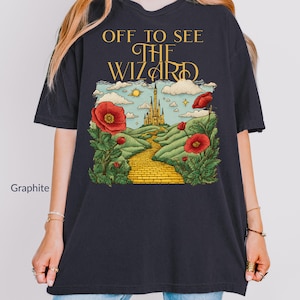 以下が含まれることがあります： 「OFF TO SEE THE WIZARD」の金色の文字が入ったグラファイトカラーのTシャツ。 黄色のレンガの道、赤いポピー、緑の丘の上の城が描かれたグラフィック。 デザインは、おとぎ話のようなテーマを持っています。