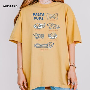 May include: Mustard-colored t-shirt with the text "PASTA PUPS" and illustrations of dogs resembling pasta shapes: farfalle, tortellini, pappardelle, rotini, orecchiette, and linguine. The shirt is a relaxed fit.