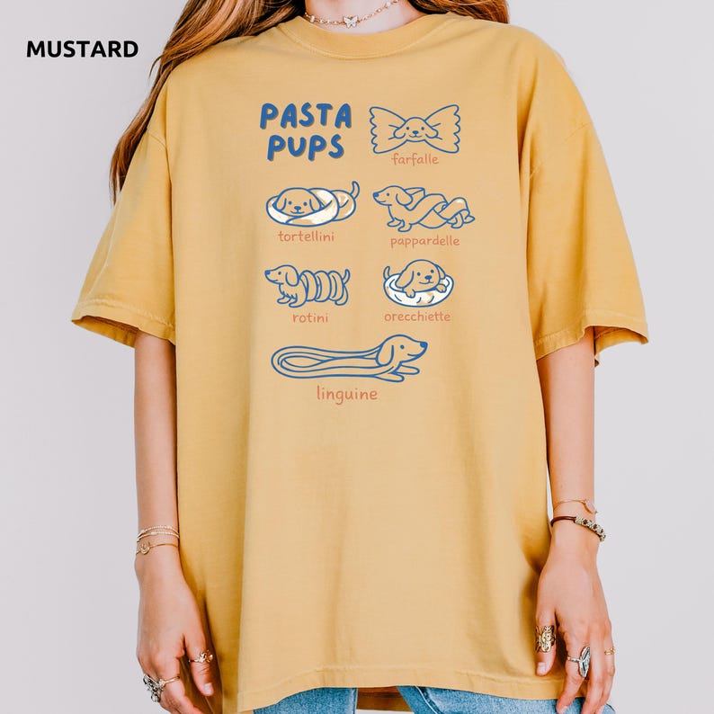 May include: Mustard-colored t-shirt with the text "PASTA PUPS" and illustrations of dogs resembling pasta shapes: farfalle, tortellini, pappardelle, rotini, orecchiette, and linguine. The shirt is a relaxed fit.